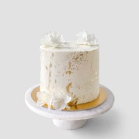The Design Your Own Cake by Jenna Rae Cakes features white frosting with gold splatters and delicate white flowers, presented on a round marble stand against a light gray background.