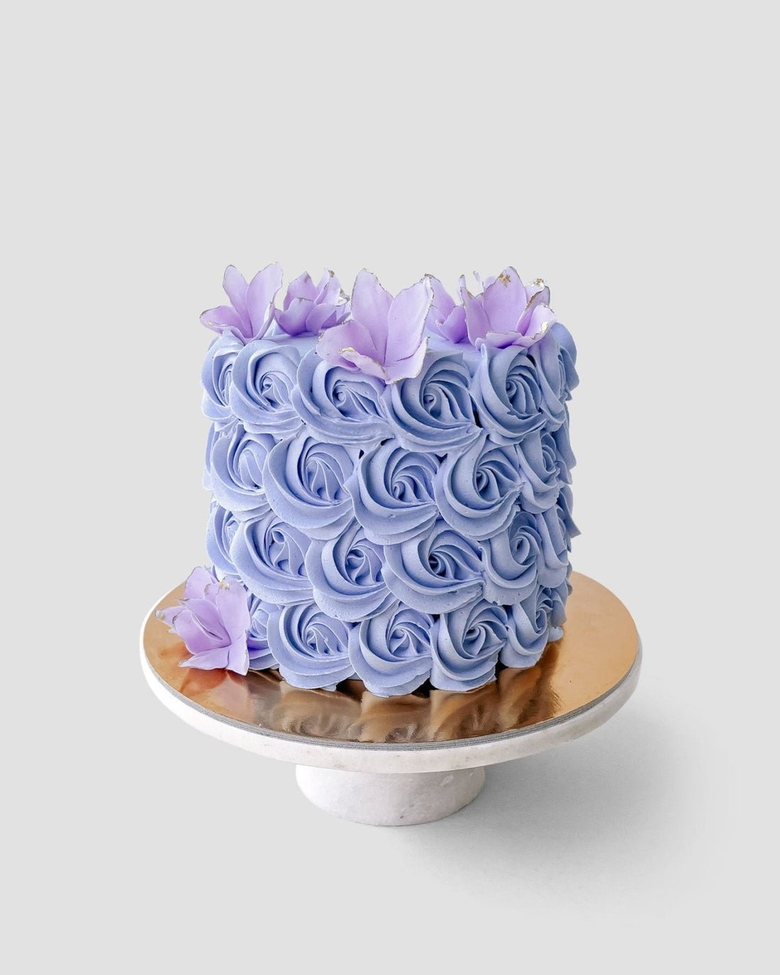 The Design Your Own Cake by Jenna Rae Cakes features a round design with intricate lavender buttercream rosettes and purple flowers, displayed on a marble cake stand against a plain light gray background.