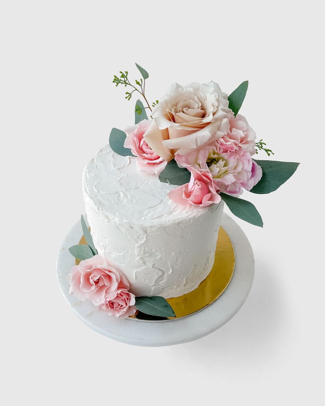 The Fresh Floral Buttercream Cake by Jenna Rae Cakes features pink and cream flowers with green leaves on white frosting, presented on a round white marble stand with a gold base against a light background.