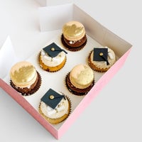 Grad Works Cupcakes by Jenna Rae Cakes: a pink box with six decorated cupcakes, three topped with gold macarons and three adorned with edible graduation caps and diplomas, arranged neatly on a light background.