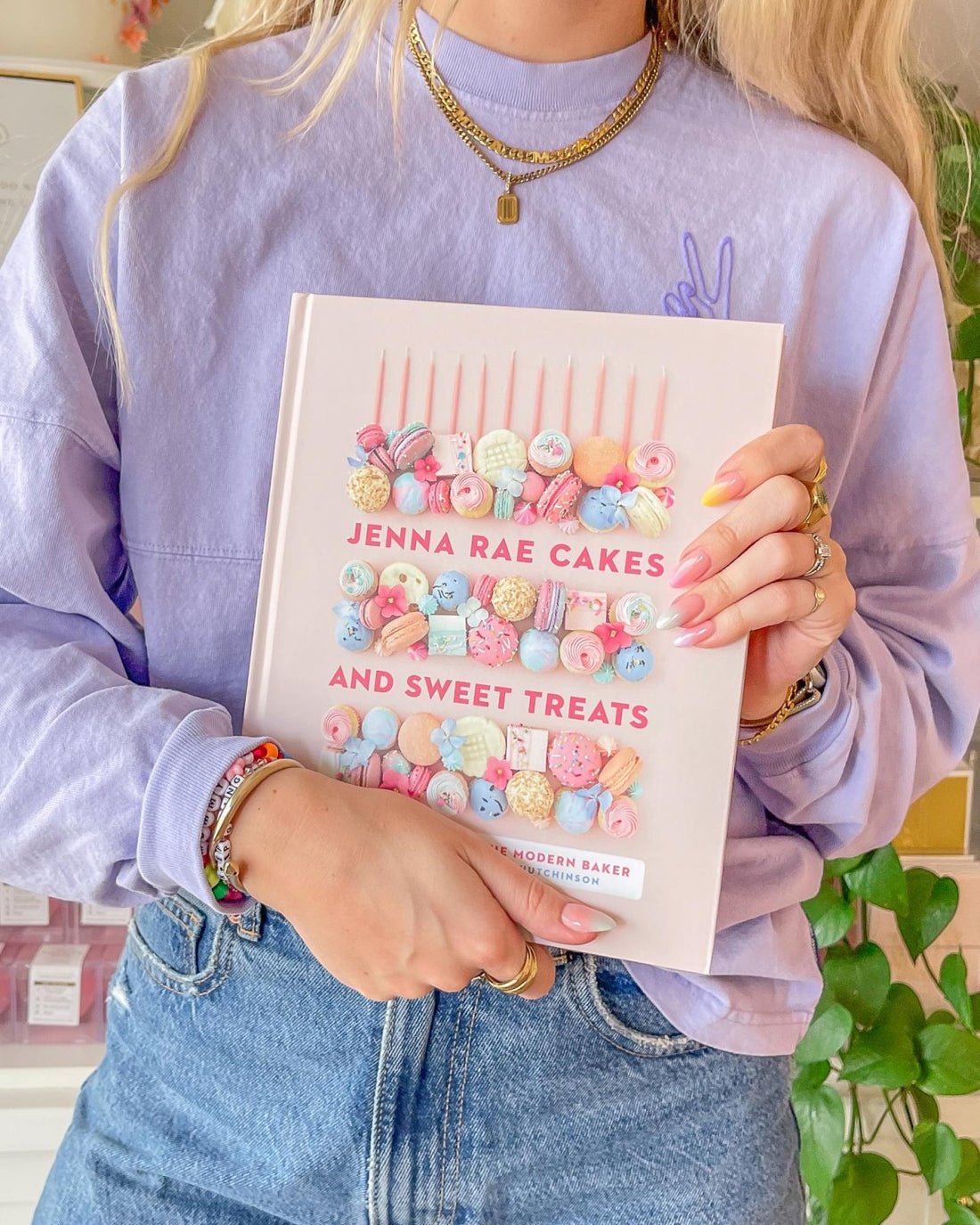 A woman in a lavender sweater and jeans holds the Jenna Rae Cakes and Sweet Treats Cookbook by Jenna Rae Cakes, featuring a pastel cover with macarons and candies. She has manicured nails and layered gold jewelry.