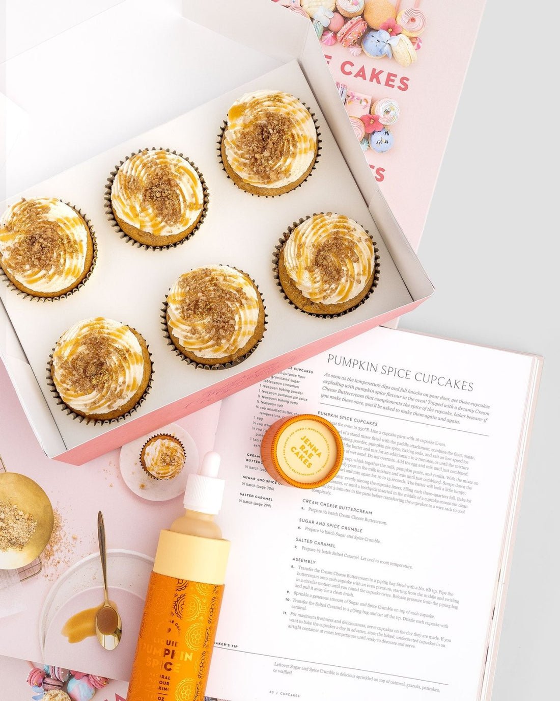 A box of six pumpkin spice cupcakes with swirled frosting sits beside the open Jenna Rae Cakes and Sweet Treats Cookbook, a piping bottle, a small container of sprinkles, and a bowl with cupcake crumbs.