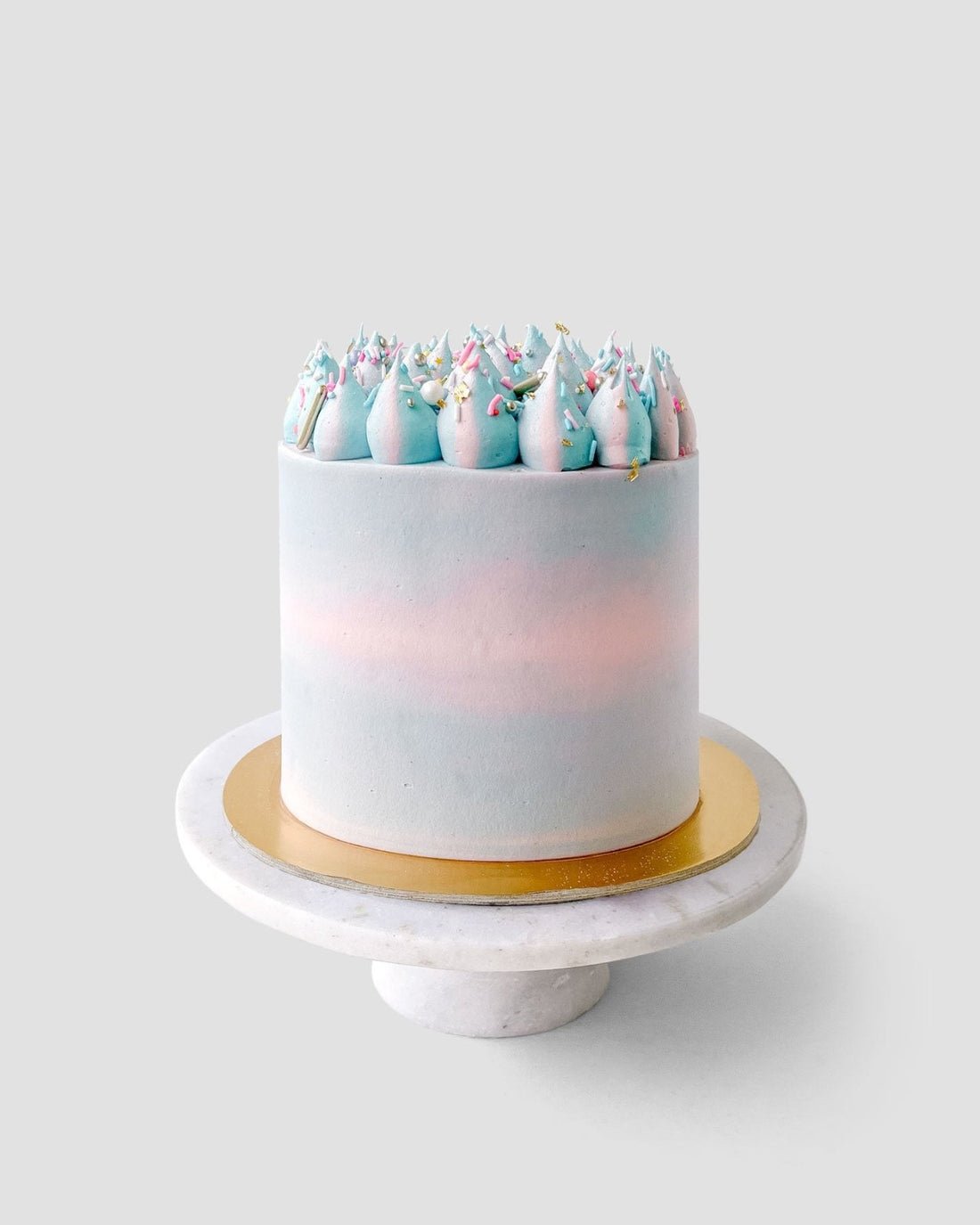 The Last Minute Cakes by Jenna Rae Cakes features a tall, round cake with smooth pastel blue and pink frosting, topped with blue and pink swirled dollops and colorful sprinkles on a white marble cake stand.