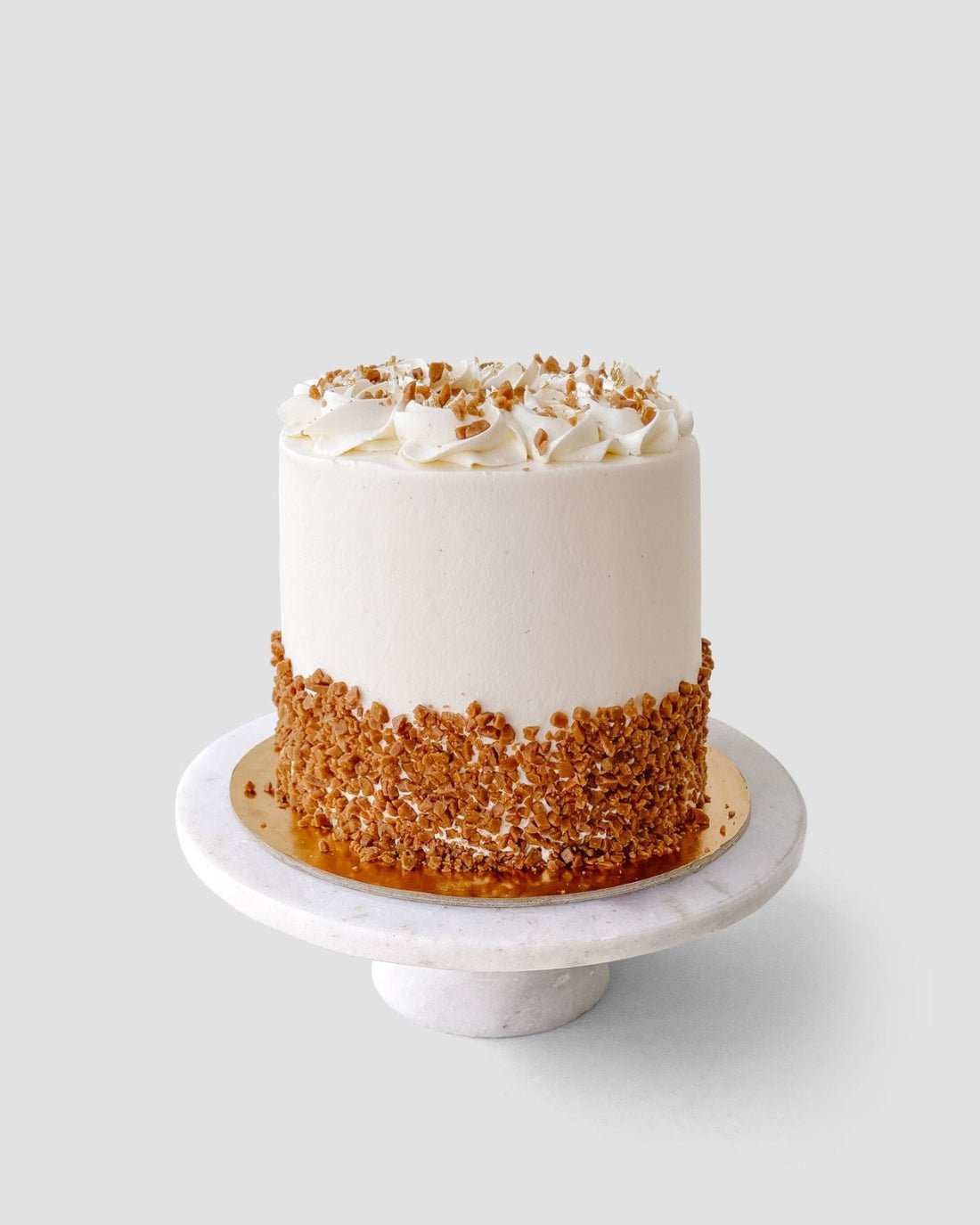 A round, white frosted cake topped with crushed nuts sits on a white marble cake stand. The lower half of the cake is coated with crushed nuts, and the top is decorated with piped swirls of frosting.