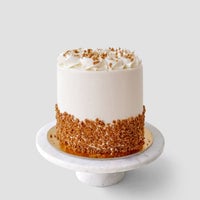 A round, white frosted cake topped with crushed nuts sits on a white marble cake stand. The lower half of the cake is coated with crushed nuts, and the top is decorated with piped swirls of frosting.