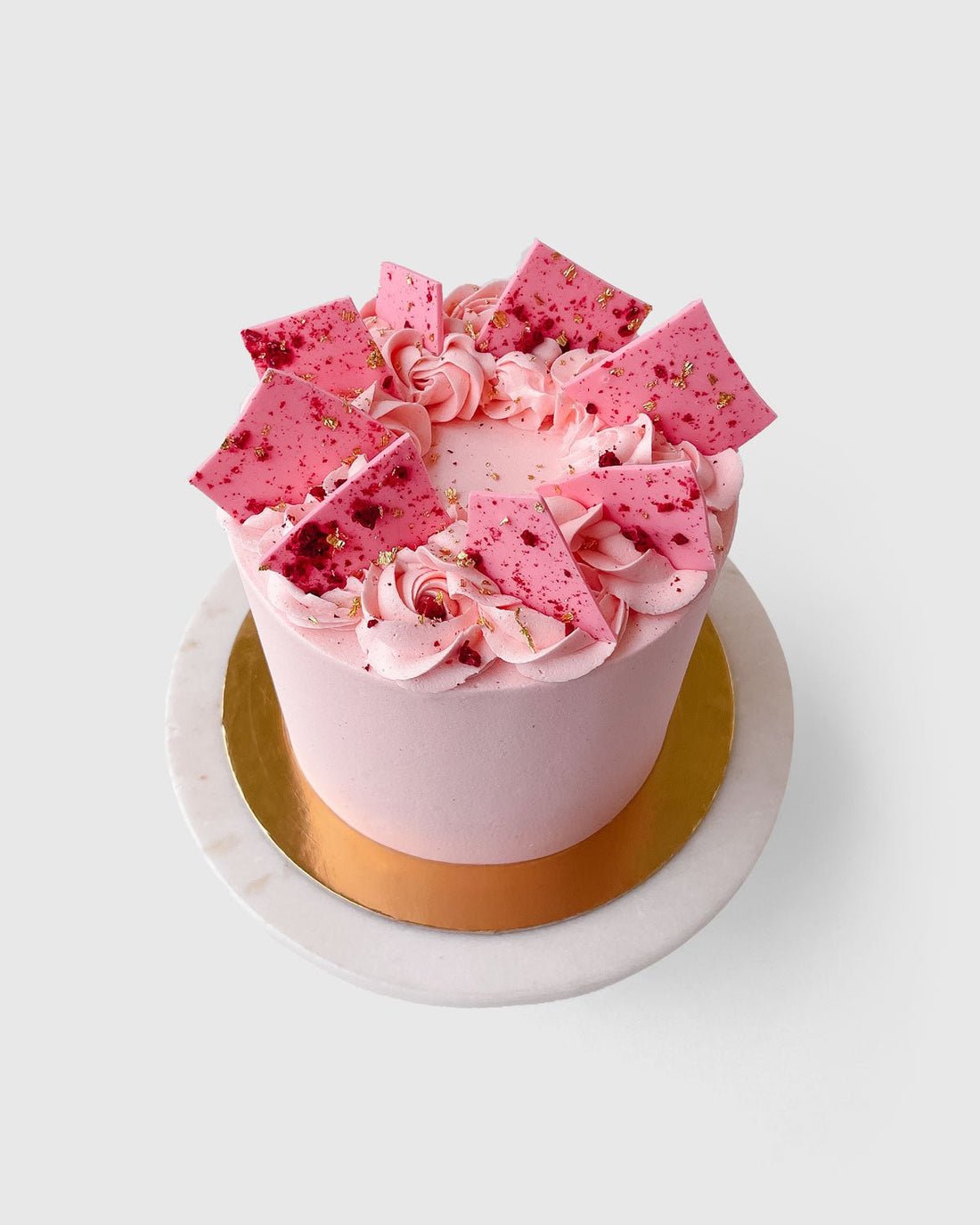 The Lemon Raspberry Cakes 101 In-Person Cake Decorating Class by Shop Jenna Rae Cakes features a round pink cake adorned with rosettes, chocolate shards, and gold sprinkles, displayed on a white marble cake stand with a gold base.