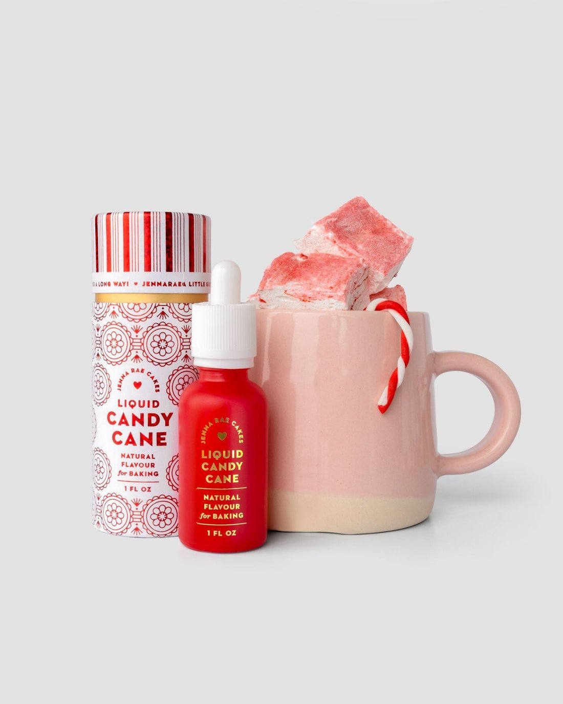 A pink mug with pink and white marshmallows and a small candy cane sits beside a Jenna Rae Cakes Liquid Candy Cane dropper bottle and matching cylindrical container.