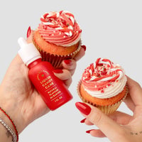 Two hands with red nails hold frosted red and white cupcakes; one hand also holds a Jenna Rae Cakes Liquid Candy Cane dropper bottle, showing it can be used for decorating or flavoring desserts.