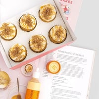 A white box of six frosted cupcakes sits next to an open cookbook displaying a recipe, with Jenna Rae Cakes Liquid Pumpkin Spice, a small container, and scattered sprinkles nearby on a light surface.