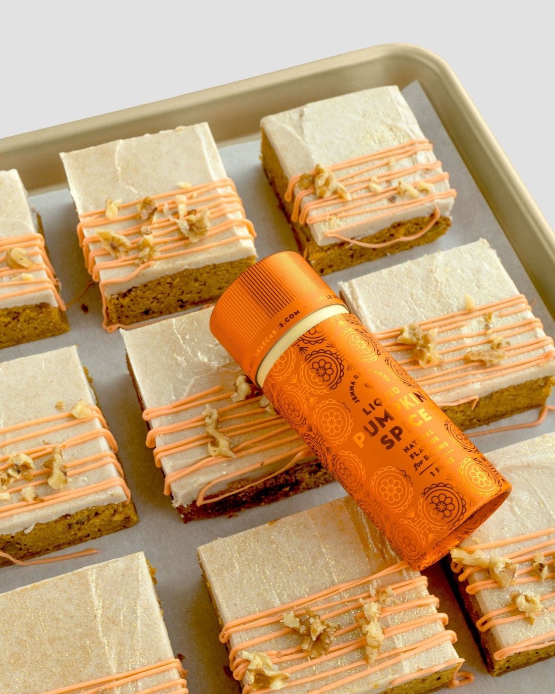 A baking sheet with frosted pumpkin spice dessert bars, topped with orange drizzle and chopped nuts, features a Jenna Rae Cakes Liquid Pumpkin Spice container resting diagonally on top of the bars.