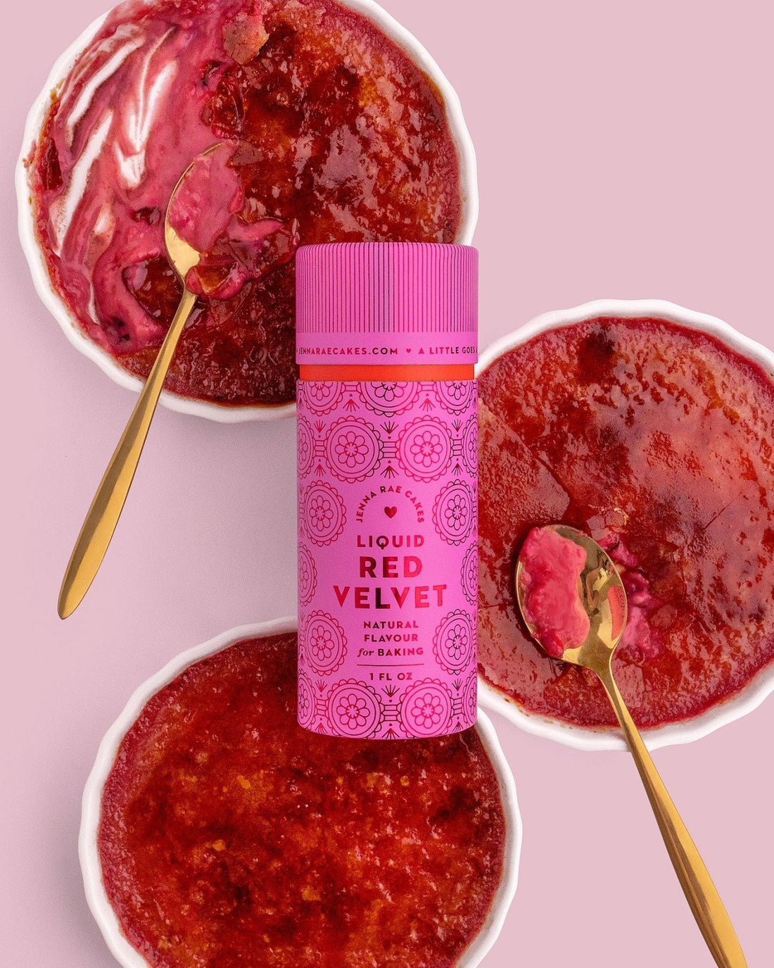 Three ramekins of red velvet crème brûlée with caramelized tops and gold spoons are arranged around a pink bottle of Jenna Rae Cakes Liquid Red Velvet natural flavoring on a pink background.