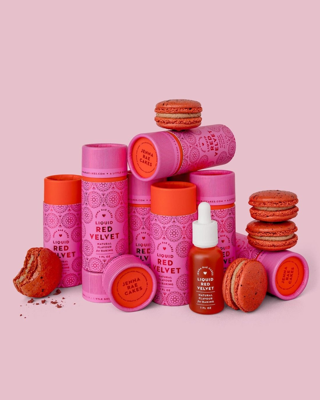 Jenna Rae Cakes Liquid Red Velvet tubes are stacked with whole and broken red velvet macarons and a dropper bottle, all on a pink background.