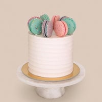 The Macaron Crown Cake by Jenna Rae Cakes features a white frosted finish topped with colorful macarons, displayed on a gold cake board and white marble stand, set against a plain beige background.