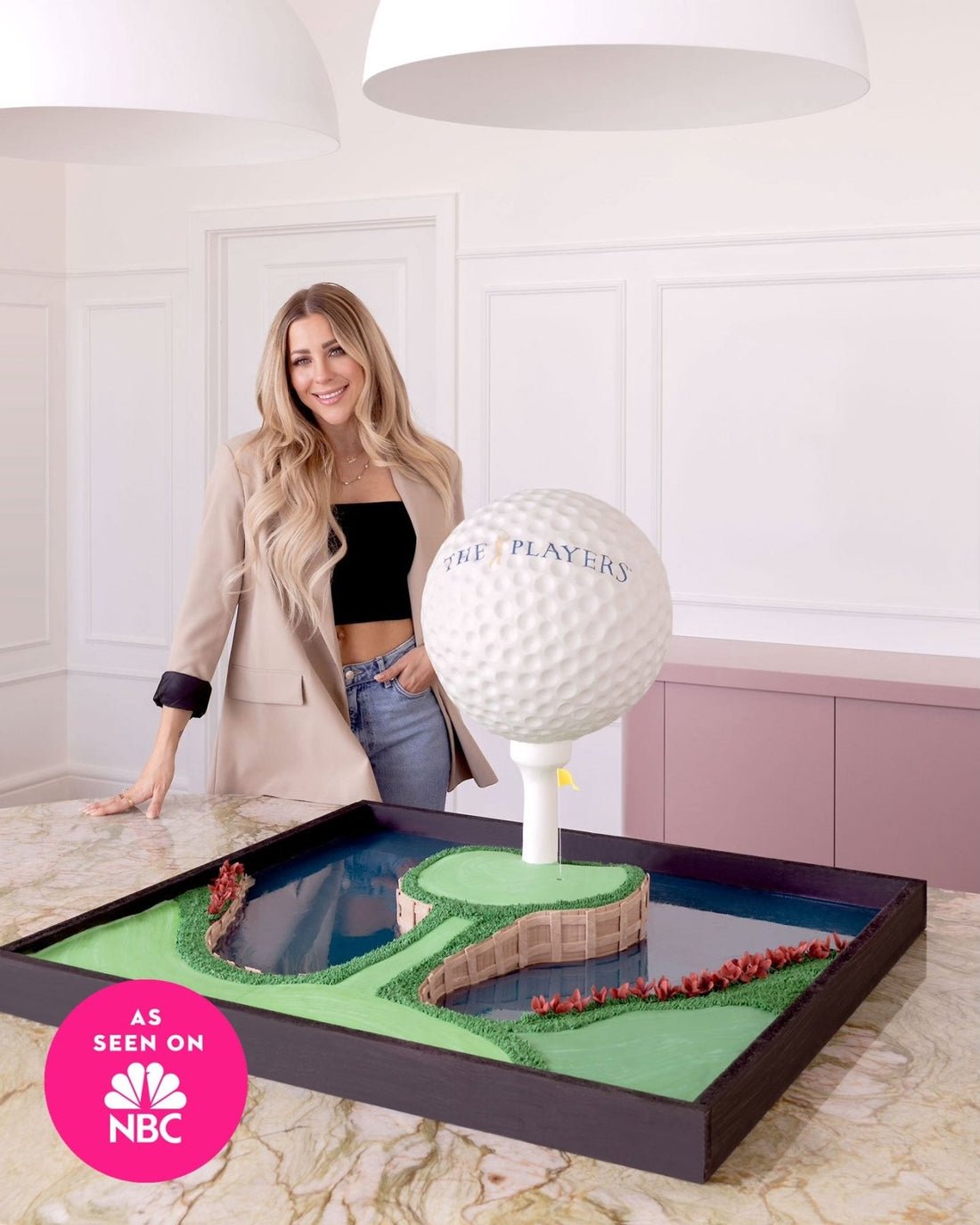 A smiling woman stands beside a large, detailed cake shaped like a golf green with a water hazard and an oversized golf ball labeled “The Players.” The cake sits on a counter, with an As Seen On NBC badge on the image.