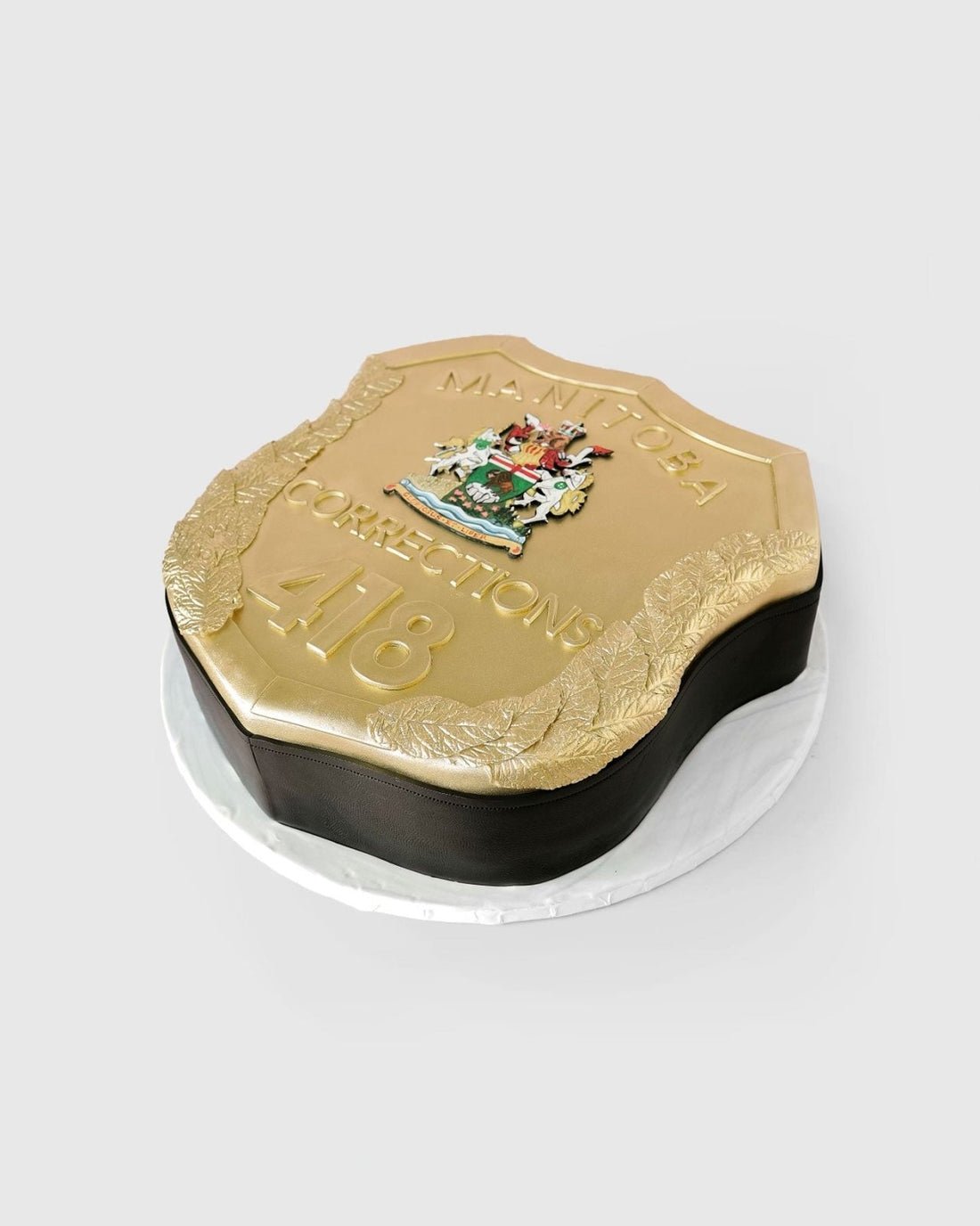 A cake shaped like a gold Manitoba Corrections badge, featuring the Manitoba coat of arms, the numbers “418,” and decorative gold detailing, displayed on a white round base.