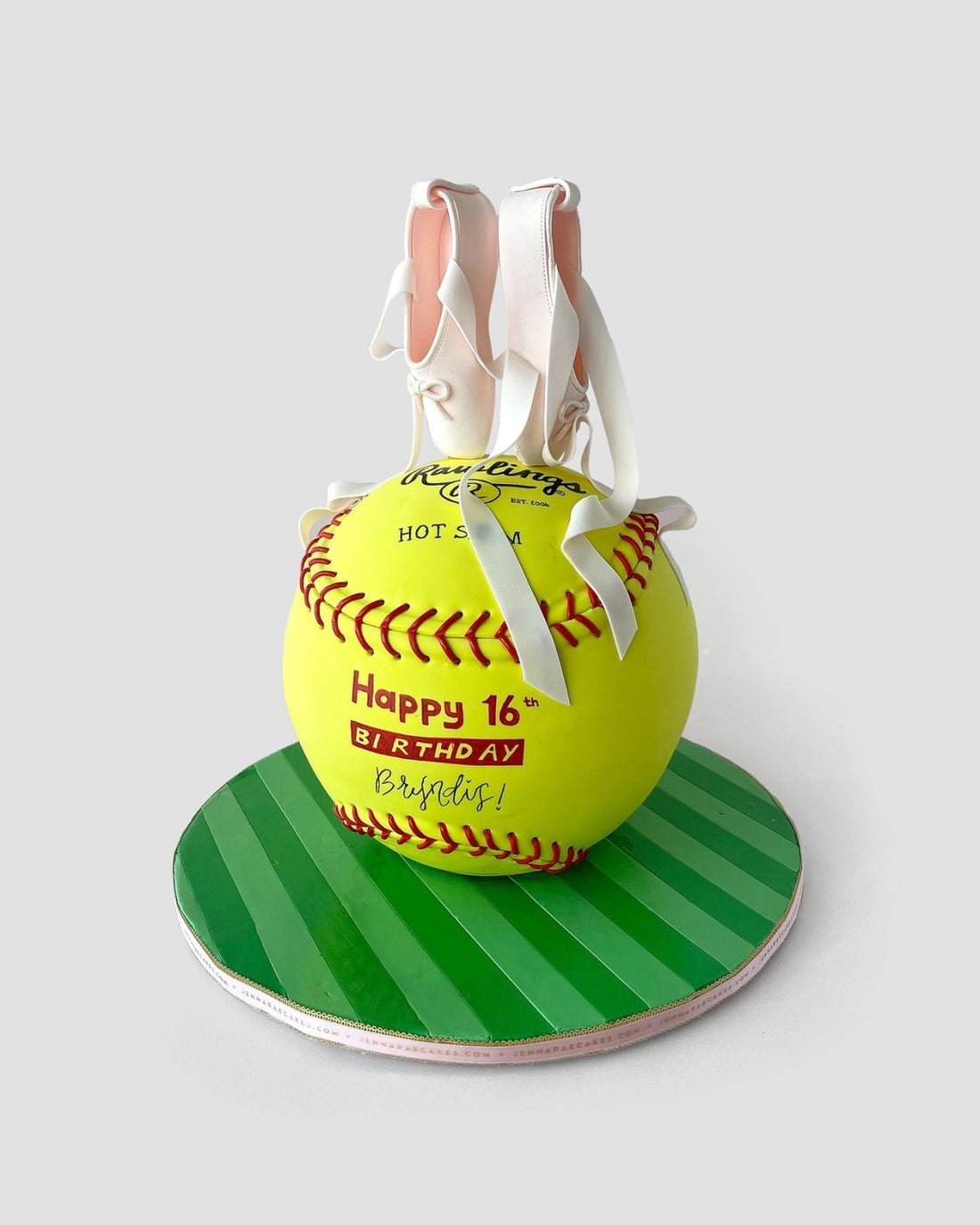 A round cake decorated as a yellow softball with red stitching sits on a green striped base. White fondant ballet slippers with pink accents rest on top. The cake reads Happy 16th Birthday Bristol! in red and yellow.