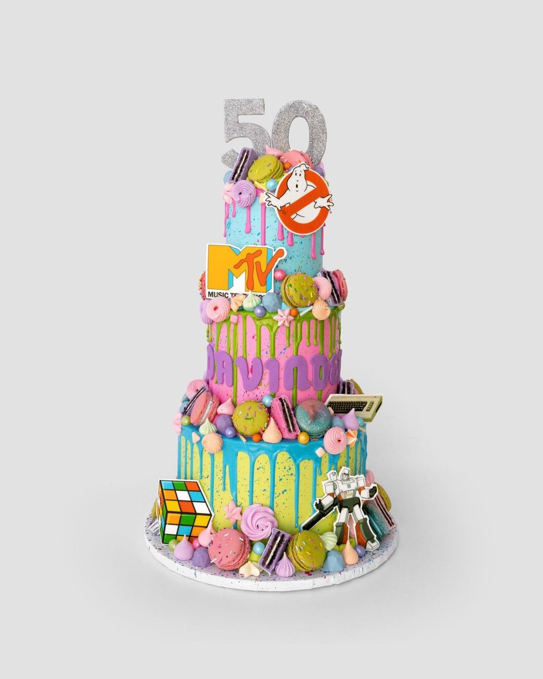 The One-of-a-Kind Cake by Jenna Rae Cakes is a vibrant three-tier treat adorned with pastel icing, macarons, and 1980s-themed toppers—Rubiks Cube, cassette tape, MTV logo, Ghostbusters sign, and the number 50.