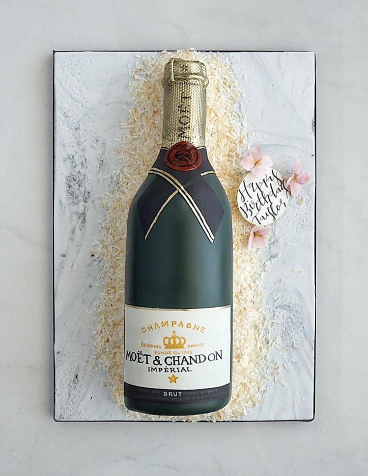 A cake decorated to look like a Moët & Chandon champagne bottle, lying on a white surface with shredded white decorations and a small note reading Happy Birthday Taylor beside pink flowers.