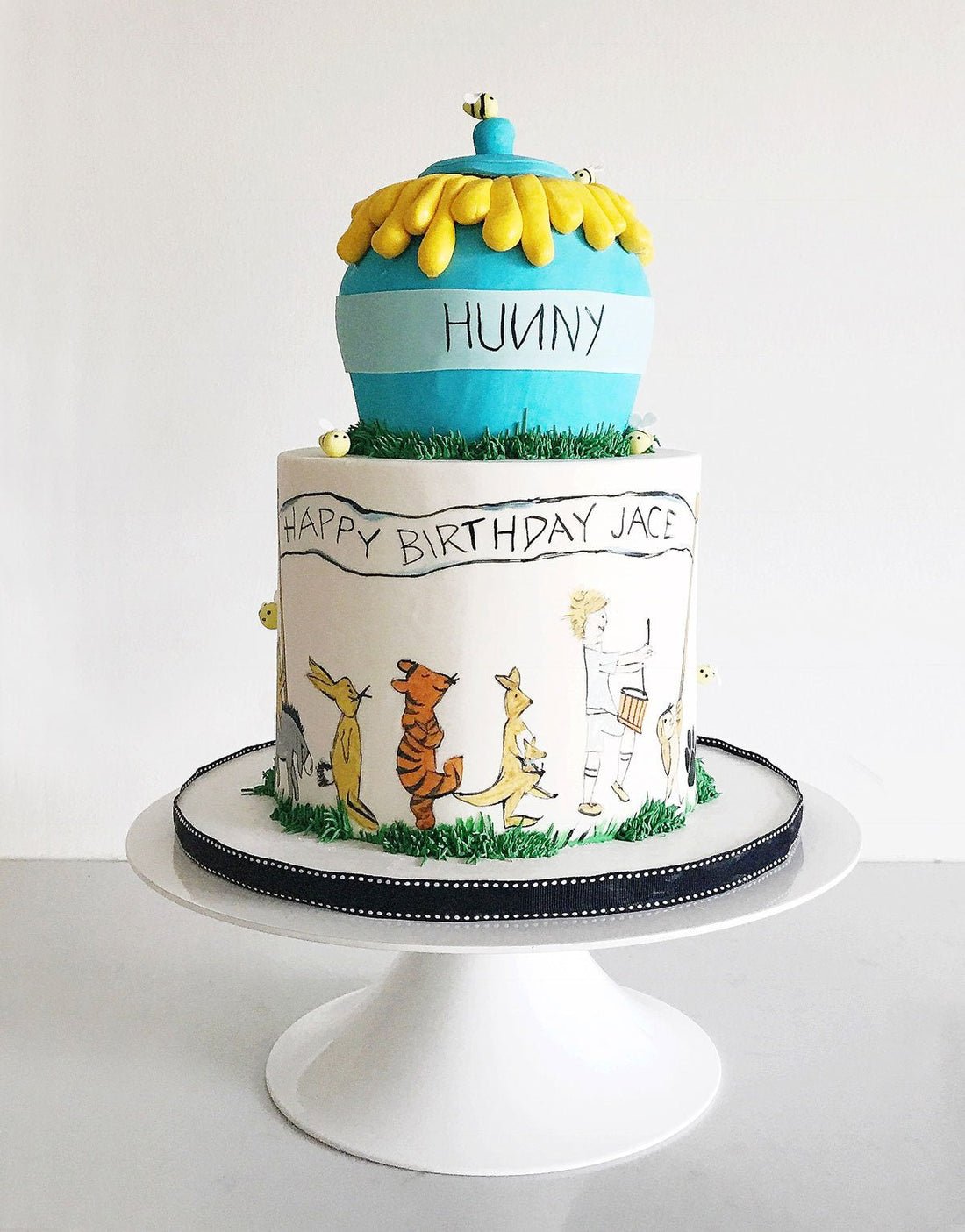 Two-tier birthday cake featuring a blue honey pot on top and drawings of Winnie the Pooh characters around the lower tier, with Happy Birthday Jace written on the side. The cake sits on a white stand.
