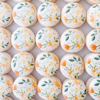 Neatly arranged on a light surface, Jenna Rae Cakes Printed Macarons feature white tops adorned with hand-painted orange flowers and green leaves.