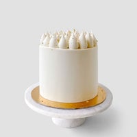 The Red Velvet Cake by Jenna Rae Cakes features white frosting with piped dollops and gold flakes, presented on a white marble cake stand with a gold base against a light gray background.