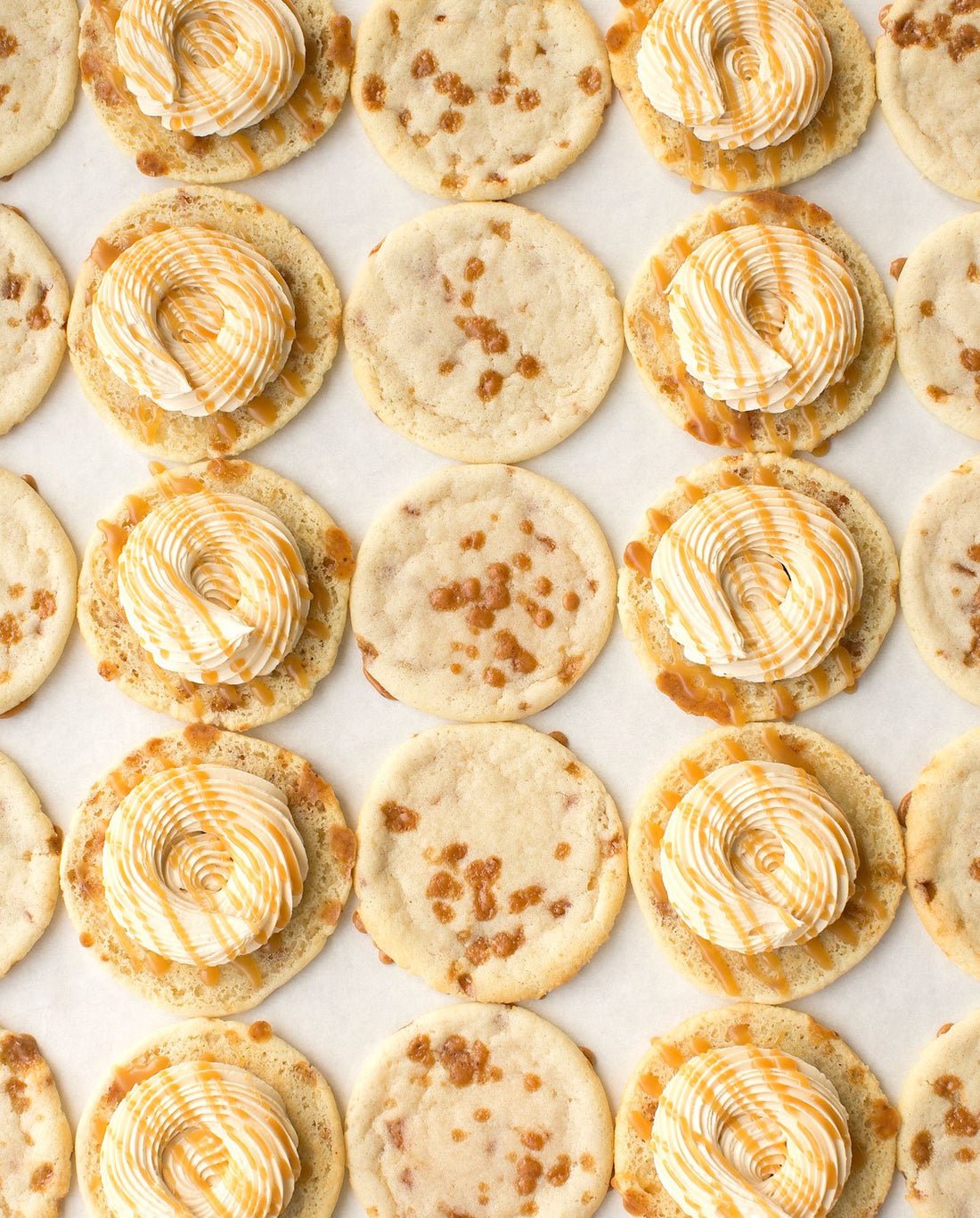 Jenna Rae Cakes Regular Cookie Sandwiches are displayed on parchment paper, featuring plain cookies, some with caramel bits, and others topped with swirled white and caramel-colored frosting.