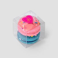 Jenna Rae Cakes’ Signature 2-Macaron Box features a clear plastic container with pink and blue macarons stacked inside, topped with colorful sprinkles and a pink heart sticker, set against a light gray background.