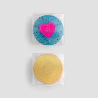 Two round soaps from Jenna Rae Cakes Signature Single Macaron Box in clear packaging on a light grey background. The top soap is blue with dark specks and a pink heart label, and the bottom is yellow with an embossed logo.