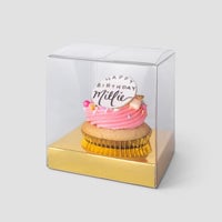 A Jenna Rae Cakes Single Cupcake Box featuring a pink frosted cupcake with a Happy Birthday Millie topper is showcased in a clear plastic box with a gold base, set against a light gray background.