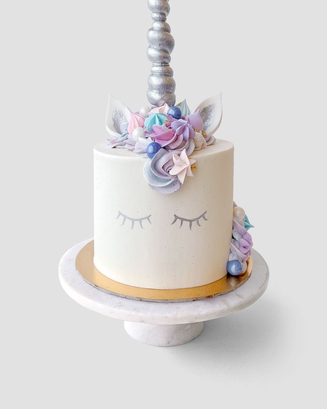 The Unicorn Cake by Jenna Rae Cakes features white frosting, closed eyes on the side, pastel flowers and ears on top, plus a silver unicorn horn, beautifully presented on a round marble cake stand.