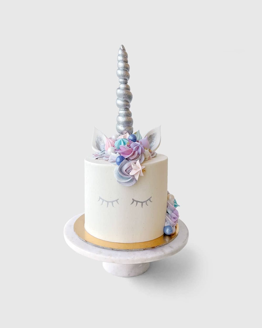 Join the Unicorn In-Person Cake Decorating Class by Shop Jenna Rae Cakes to create a white unicorn cake with closed eyes, silver horn, pastel floral mane, and ears, all beautifully displayed on a marble stand against a simple backdrop.