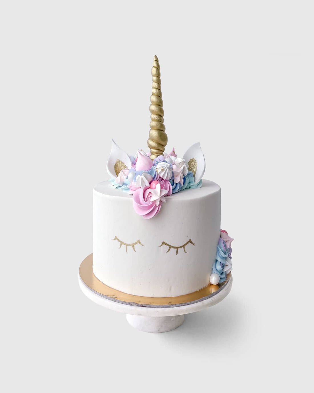 The Unicorn Cake by Jenna Rae Cakes features white frosting, a gold horn, closed eyes, pastel flowers, and ears on top, displayed on a marble cake stand against a plain background.
