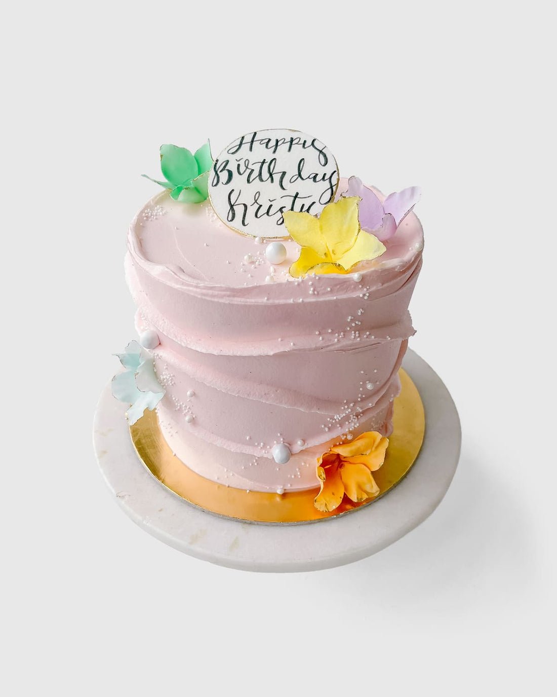 The Waves and Pearls cake by Jenna Rae Cakes features pink frosting, colorful edible butterflies, white sprinkles, a round Happy Birthday topper, and sits on a gold and white cake board.