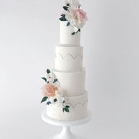A four-tiered white wedding cake decorated with delicate pastel flowers and greenery, featuring subtle gold accents and pearl-like borders, displayed on a white stand against a plain background.