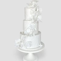A three-tiered white cake decorated with delicate, white sugar flowers cascading down the side, displayed on a simple white cake stand against a plain light gray background.