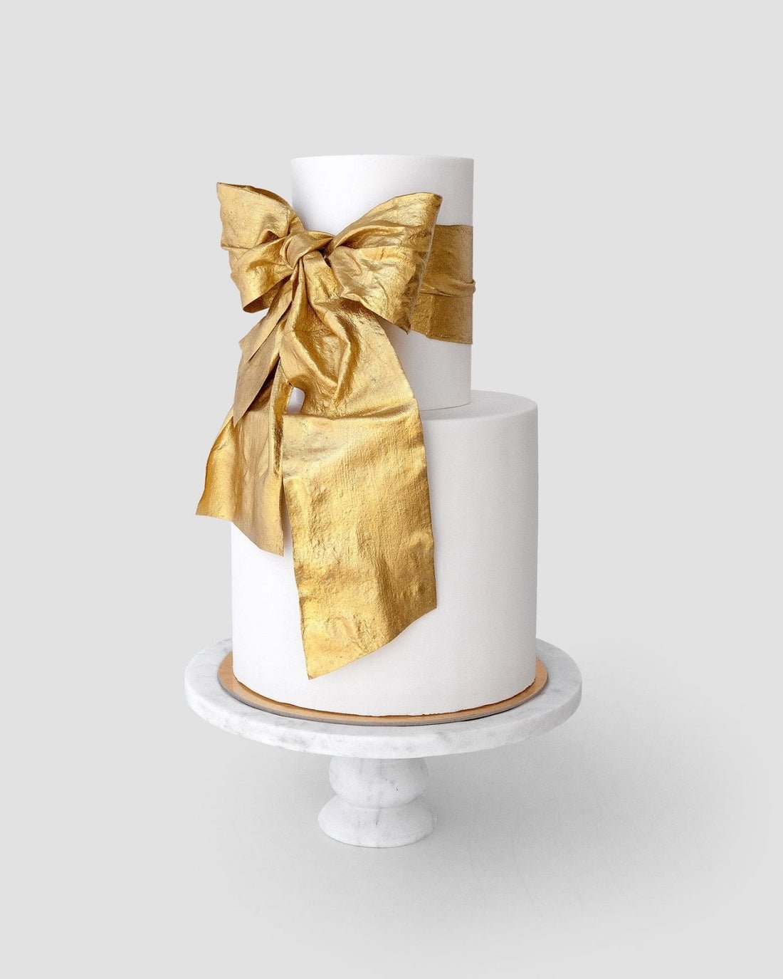 Jenna Rae Cakes’ “Wedding Cakes Under $1,000”: A two-tier white cake on a marble stand, topped with a statement metallic gold bow.