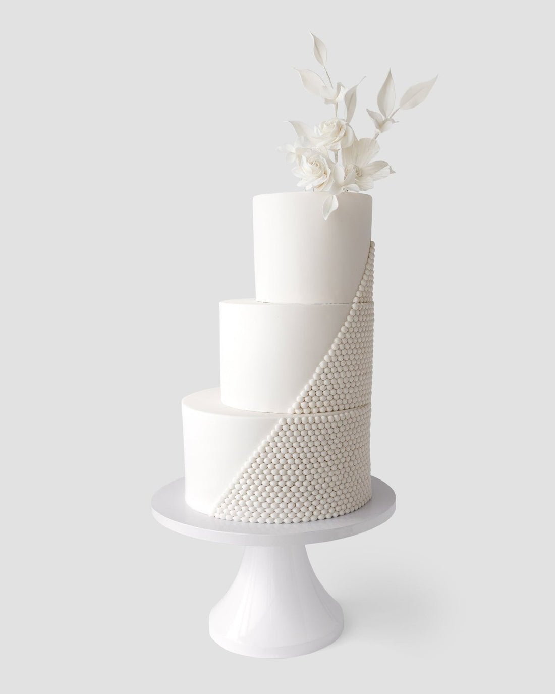 The Wedding Cakes Under $1,000 by Jenna Rae Cakes features a three-tiered white fondant cake with a diagonal cascade of pearl-like beads and white flowers with leaves, elegantly displayed on a white pedestal stand.