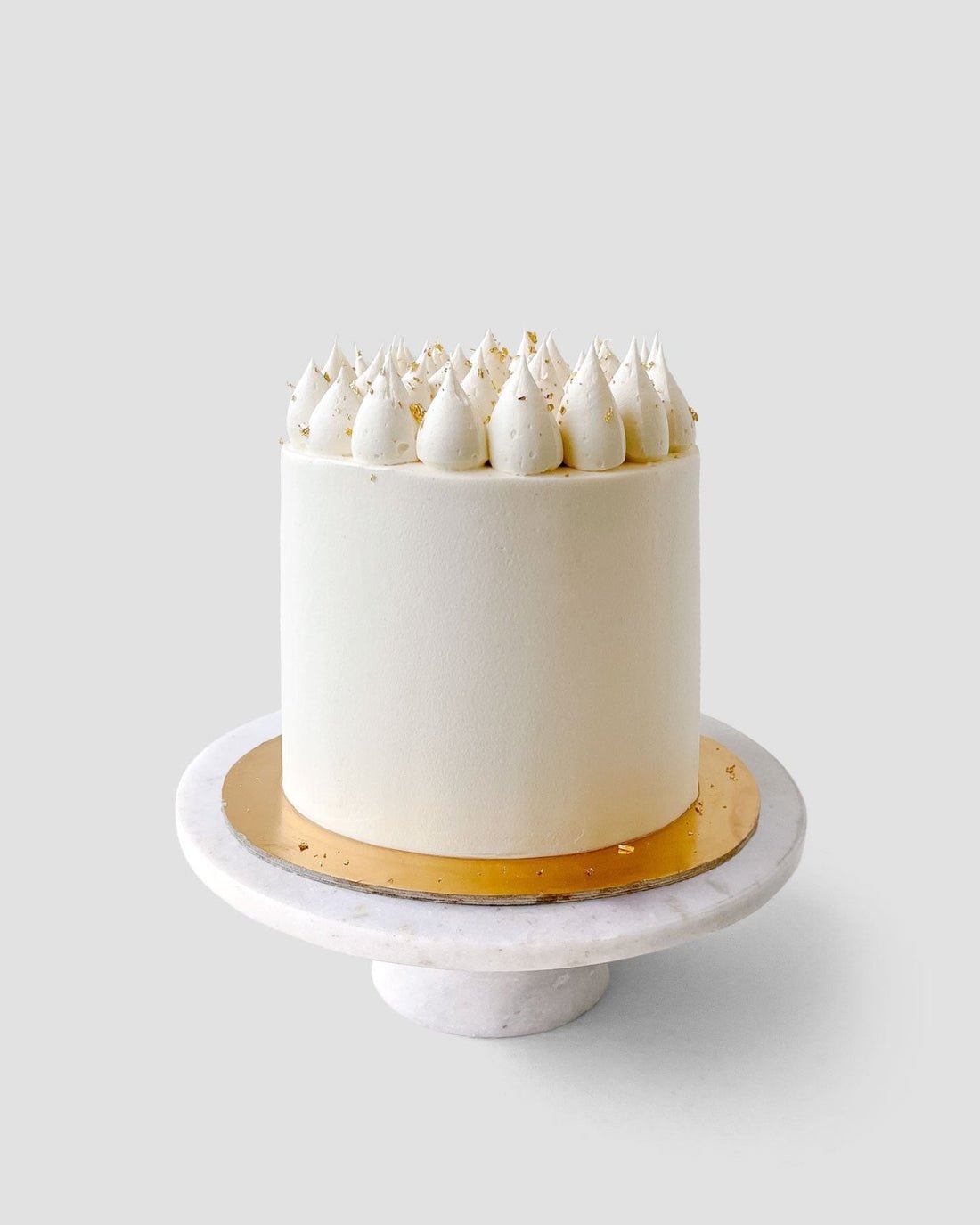 A Wedding Cake from Jenna Rae Cakes under $200 features white frosting, whipped cream dollops, and gold sprinkles on a gold cake board and white marble stand against a light gray background.