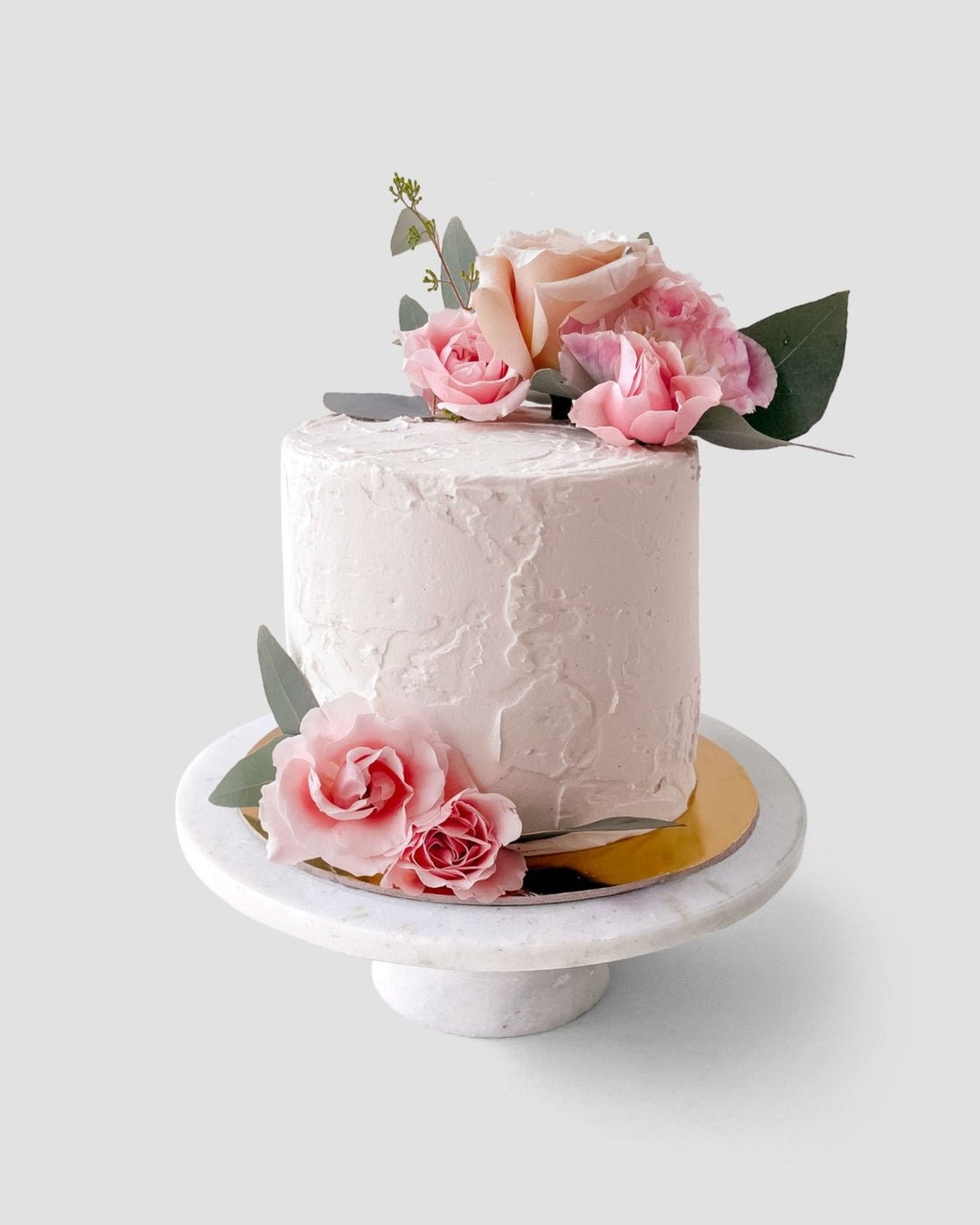 The Jenna Rae Cakes Wedding Cakes Under $200 features a white frosted cake on a marble stand, adorned with pink and peach roses and green leaves.