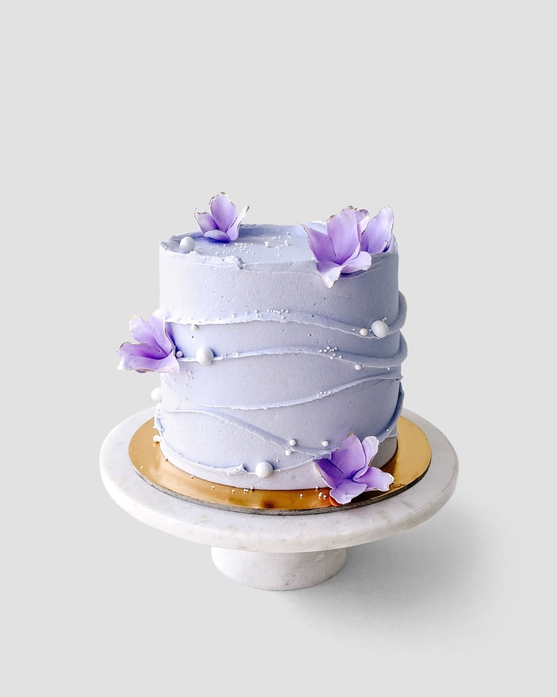 Discover Jenna Rae Cakes Wedding Cakes Under $200: a round, light purple cake adorned with wavy icing lines, white sugar pearls, and purple sugar flowers—beautifully displayed on a marble stand with a gold base.
