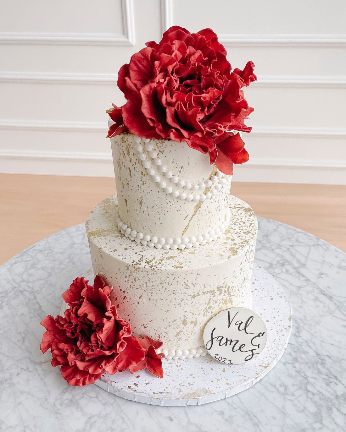 Jenna Rae Cakes’ Wedding Cakes Under $500 features a two-tier white cake with gold speckles, bold red flowers, pearl-like beads, and a custom “Val & James 2024” plaque on a marble-patterned board.