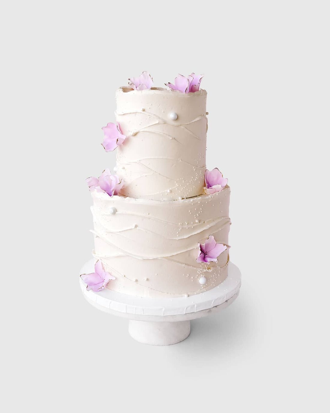 A two-tiered white frosted cake with light pink edible flowers and pearls by Jenna Rae Cakes, featured in Wedding Cakes Under $500, displayed on a white stand against a plain light background.