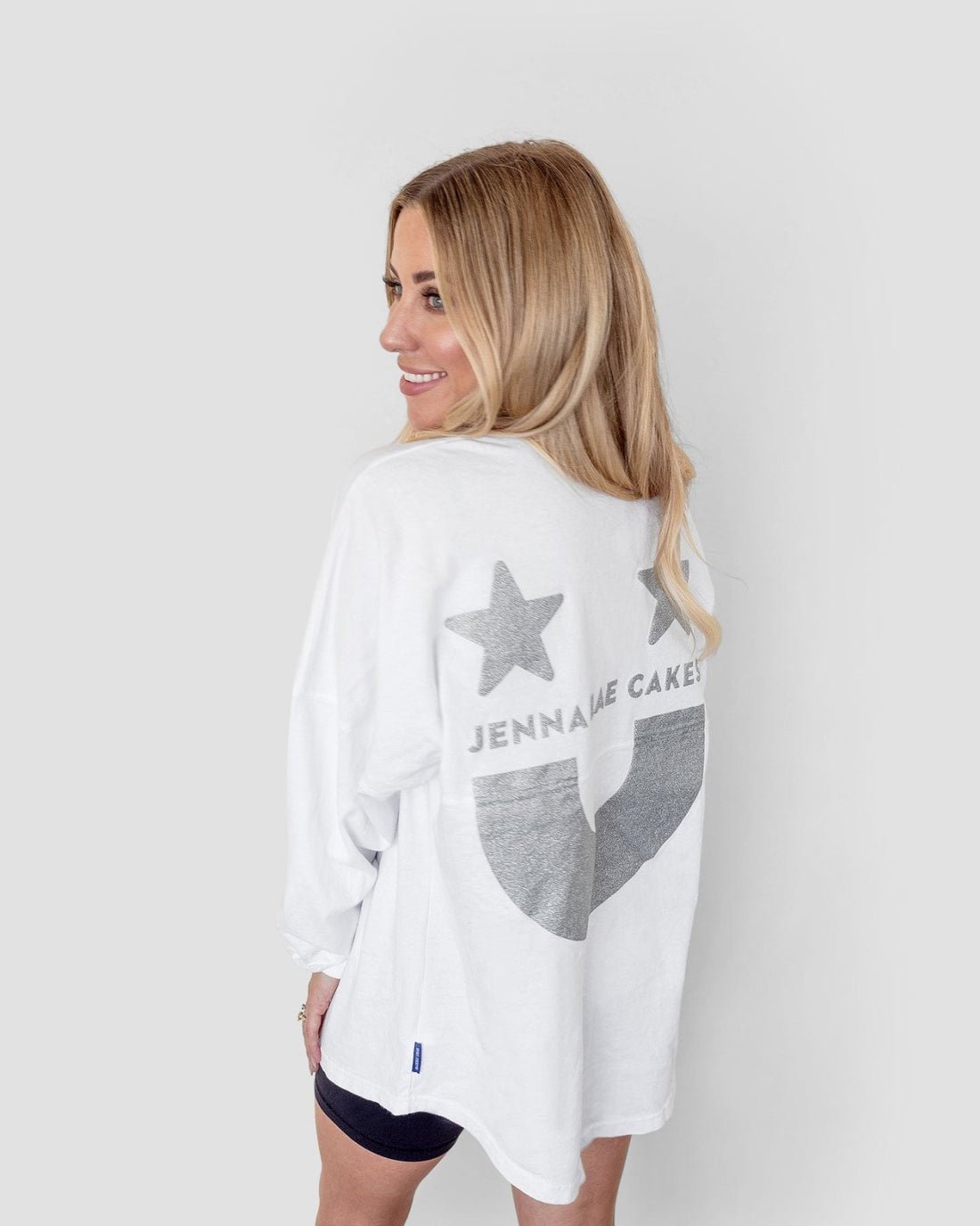 A woman with long blonde hair smiles over her shoulder, wearing the Jenna Rae Cakes White and Silver Spirit Jersey with two stars on the back, paired with black shorts.