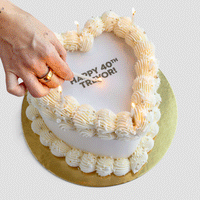 A hand lights candles on Jenna Rae Cakes’ Burn Away Heart Cake, a heart-shaped white cake with piped icing on a gold board. “HAPPY 40th TREVOR!” is written in gray letters.