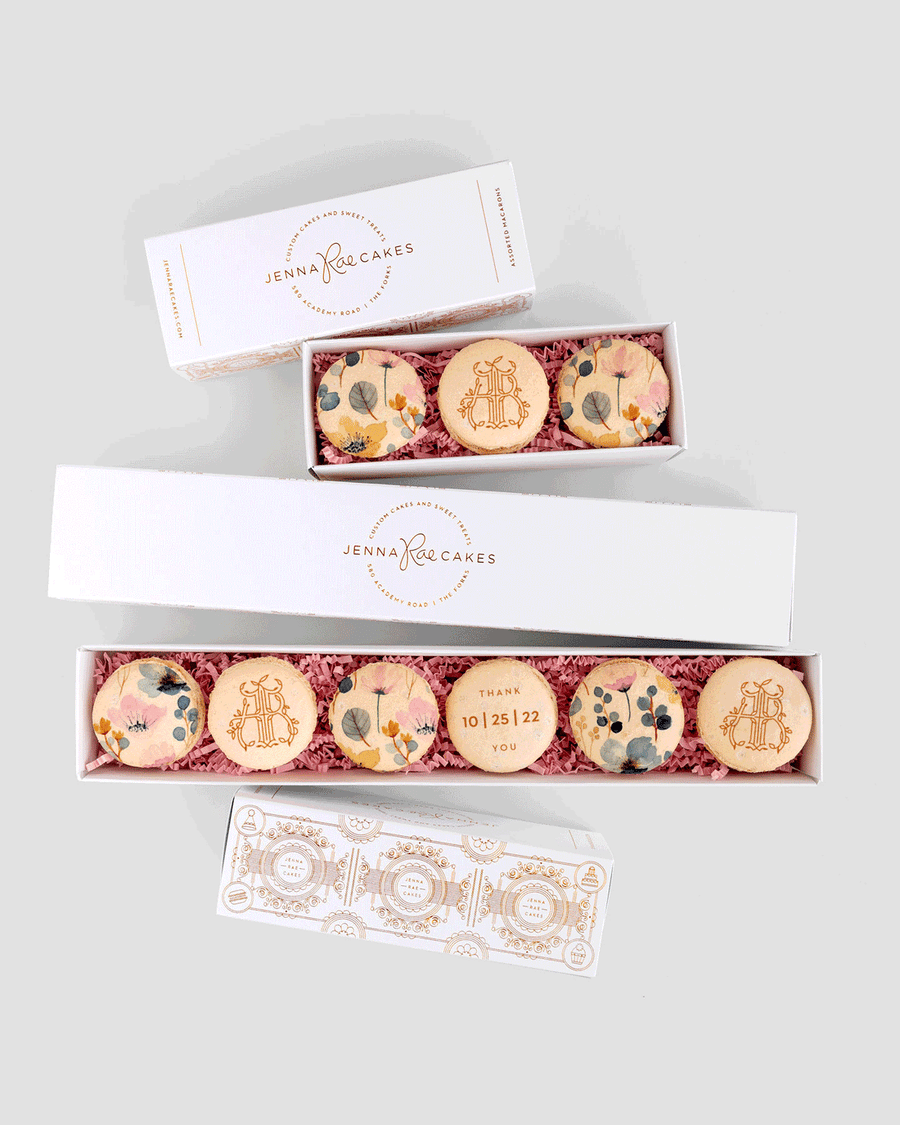 Three white Favours boxes from Jenna Rae Cakes, each holding decorated macarons with floral designs and gold initials, displayed on a light gray background. One macaron features THANK YOU 10 25 22.