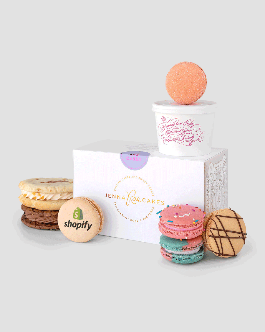 The Jenna Rae Cakes Sweet Treat Gift Box is displayed with assorted colorful macarons, a Shopify logo cookie, and a small cup topped with a pink macaron, all arranged on a light gray background.