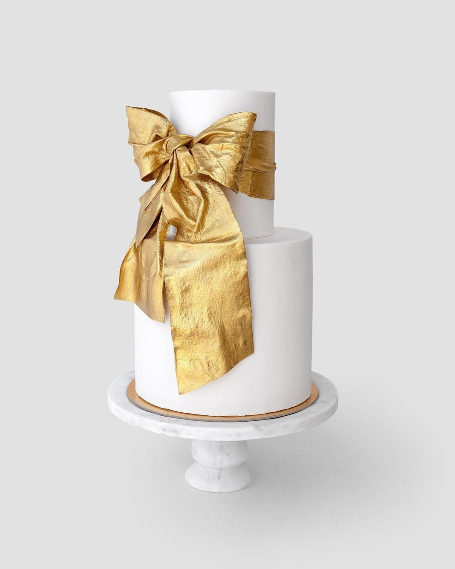 A two-tiered white cake on a marble stand, decorated with a shimmering antique gold bow made using Jenna Rae Cakes Antique Gold Edible Lustre Dust - 5g Shaker (Package of 6).