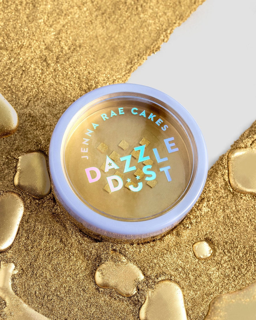 A round container from the Jenna Rae Cakes Dazzle Dust Best Seller Trio rests on a surface dusted with gold powder and liquid splatters, featuring a lavender rim and holographic text on its lid.