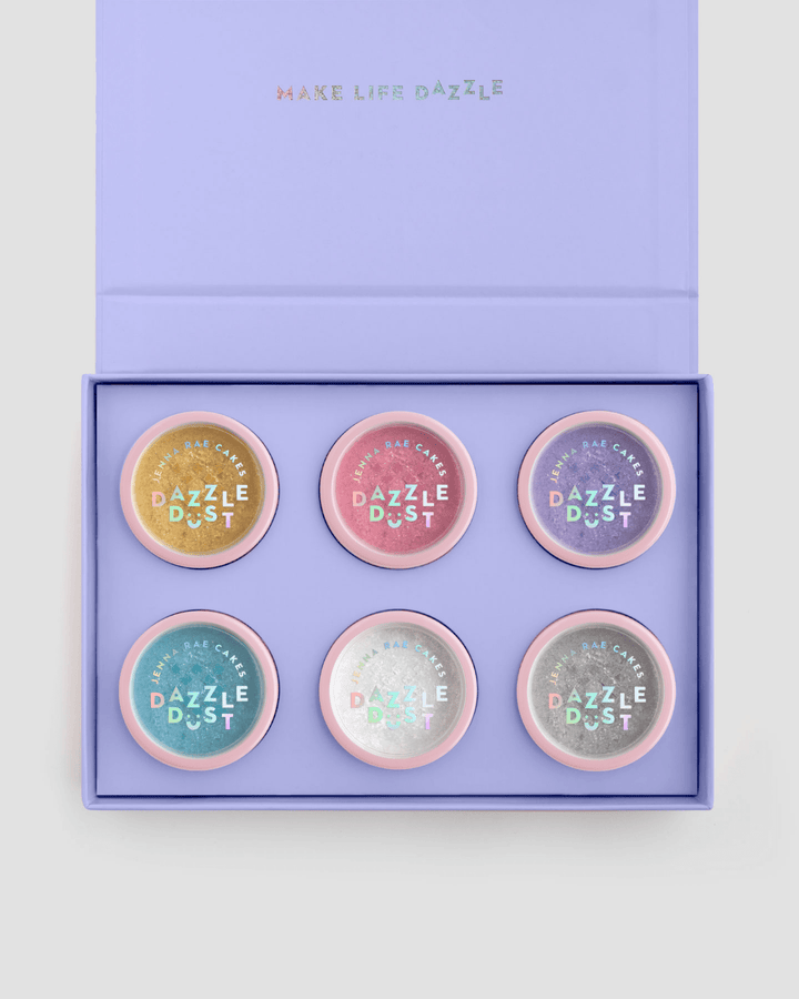 The Build Your Own Dazzle Dust Collection by Jenna Rae Cakes features six round pastel containers labeled Dazzle Dust, arranged in two rows of three inside a lavender gift box with MAKE LIFE DAZZLE on the lid.