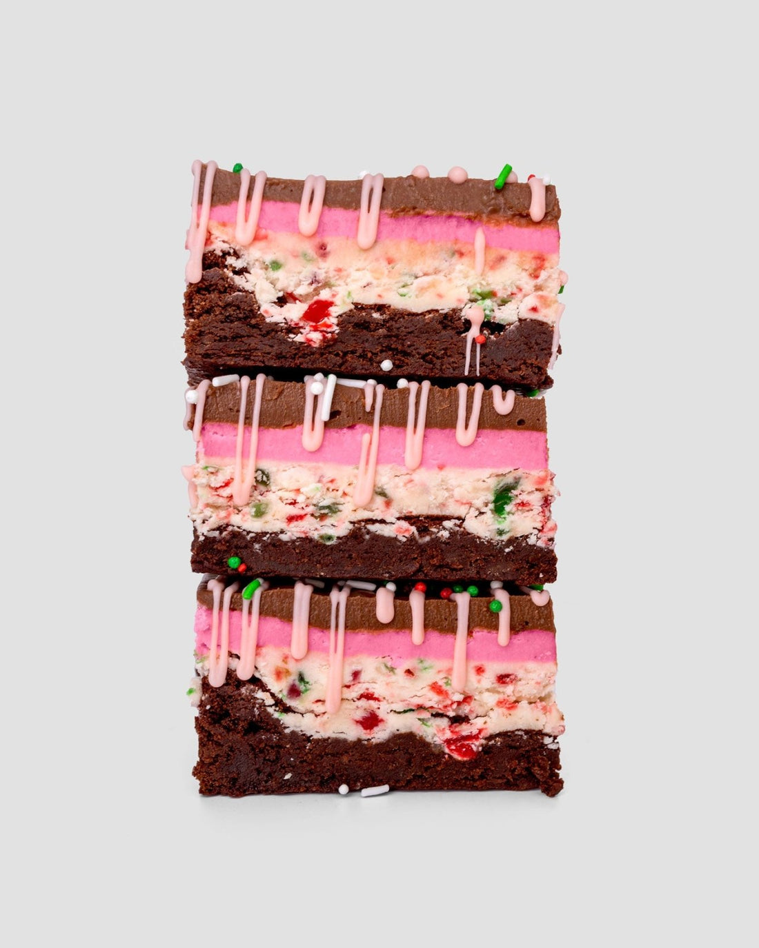 Three stacked Jenna Rae Cakes Candy Cane Brownie bars—limited edition—feature chocolate layers, pink frosting, white cream with red and green sprinkles, and a pink icing drizzle, set against a plain light background.